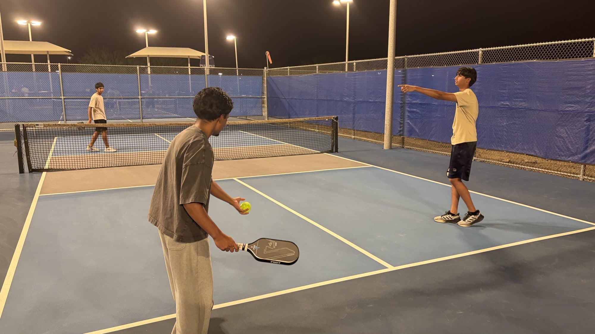 A player holding the ball and paddle at the baseline, lining up a serve while his partner signals across the court